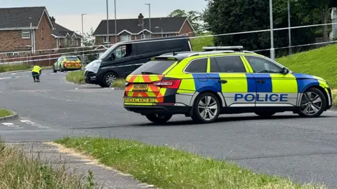 A police car parked across a road behind it there is police tape and a person in hi-vis clothing.