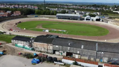 Dan Adams The Swindon Abbey Stadium seen in an aerial shot. It has a racetrack with green grass in the centre. There are grey stands for spectators to stand and sit.