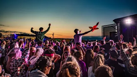 Tiree Music Festival A packed outdoor audience gathers in front of a stage as the sun sets, with people raising their hands and enjoying the live music under colourful lights.
