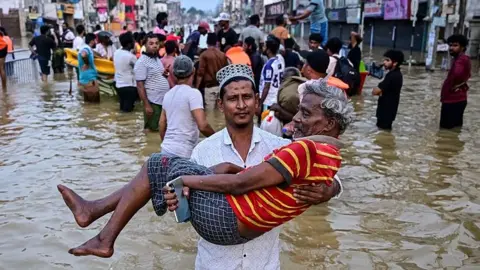 A man looking into the camera as he carries an older man in his arms, on a flooded road