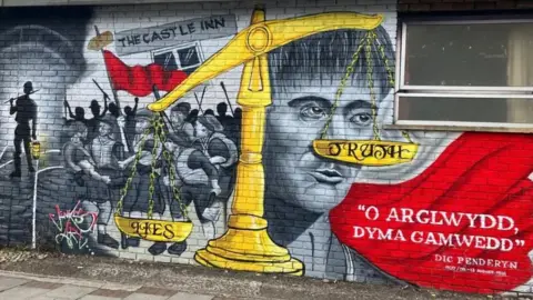 A mural painted onto an outdoor brick wall in Port Talbot, the artwork is a memorial to Dic Penderyn (Richard Lewis),  A large, yellow scale of justice is unbalanced, labeled "LIES" weighing heavily downward on the left, while the pan labeled "TRUTH" elevated on the right.  With a painting of Richard Lewis' face behind the scales.