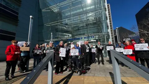 A group of men and women and a person in a wheelchair are standing outside Endeavour House holding placards saying "Don't wreck our libraries". 