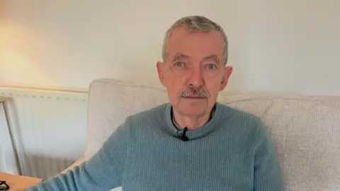 Image of a man in a blue jumper sitting in on a sofa. 