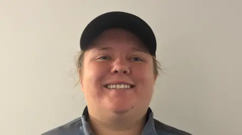 Lauren Revill stands in front of a white wall and smiles. She wears a cap with some of her hair showing from underneath. She wears a black and grey jumper with a blue top underneath.