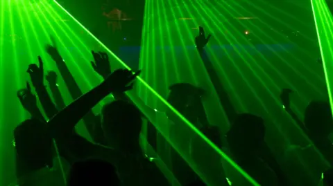 People dancing with arms aloft. They are in silhouette with green lasers shining down on them.