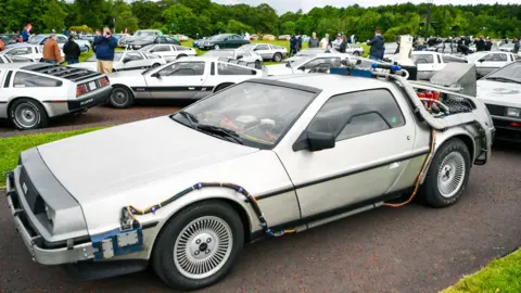 Pacemaker A Delorean car. It is silver, and there are a number of other Delorean cars in the background. 