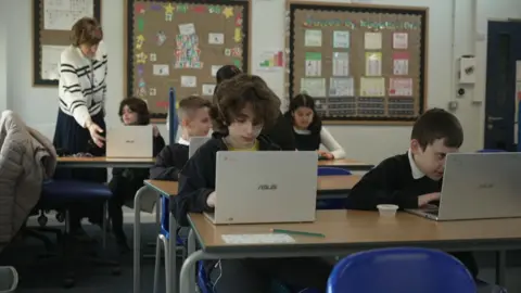 School children sitting at desks in a classroom setting with laptops.