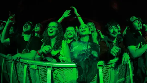 Getty Images A crowd of young men and women smile and raise their arms as they look towards a stage, shrouded in a green light