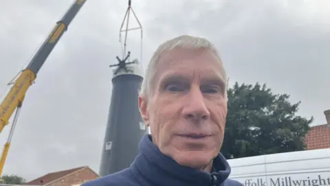 Man with grey, wearing a blue top and is standing in front of a very tall black windmill which has no sails. A yellow crane has lifted the white cap into place on the top of the windmill. There are grey skies and a tree and white van behind the man too.