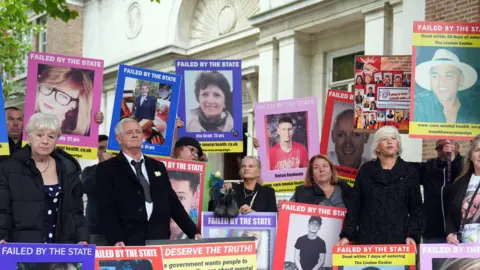 The families of those who have died while in the care of mental health services in Essex stand outside the inquiry protesting. There are around 20 people in the image which shows them standing with placards held against them and large posters of their loved ones. They show a range of faces, both men and women and young and old. One reads "failed by the state".