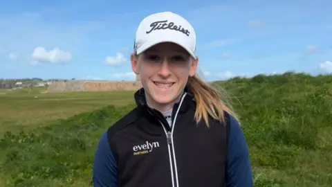 Anabelle Lucas-Villar, a young woman with light brown hair, smiles into the camera. A white golf cap shadows her eyes. She's also wearing a black gilet over a dark blue sports jumper. Behind her is a golf course, with a green mound and old fortifications.