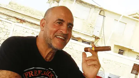 US Department of Justice Daniel Siad, a man with a shaved head and stubble, wearing a black T-shirt, sits outside in what appears to be a sunny street, smiling with a large cigar raised in his hand.