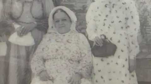 Halle Faraj A black and white image of a woman sat down on a chair. Two other women are standing on either side of her, clutching handbags, but neither woman's face is visible. The seated woman has her hair tied back and is wearing a head covering. Her dress appears pale, with a circular print.