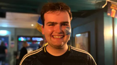Shaun McGeachan, wearing a Scotland top and with a saltire painted on his left cheek, smiles while standing inside a pub.
