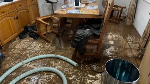 Mud is all over a kitchen floor after it was flooded. The remaining water is brown. A hosepipe is on the floor. Kitchen chairs have fallen over and are sideways on the floor. A wooden kitchen table is in the centre of the room. Place mats are on the table.