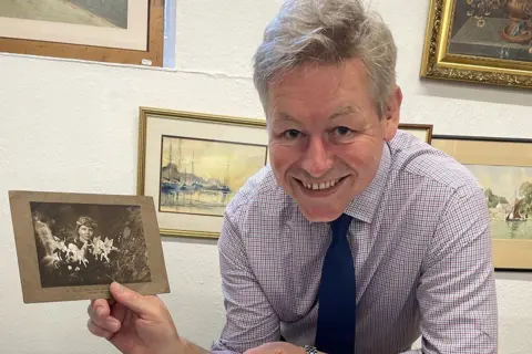 John Taylors Auction Rooms A man with grey hair, a purple and white square patterned shirt and navy tie is holding a black and white photo of a girl with fairies in front of her. Behind him are paintings on a wall.