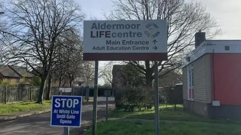 Exterior of Aldermoor Life Centre building, with trees in the background. One sign says Aldermoor Life Centre and directions to main entrance, a smaller blue sign on the left with white writing saying 'Stop at white line until gate opens'. 