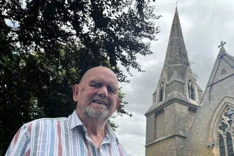 Emma Baugh/BBC John Reach has a short grey beard and is wearing a striped blue, green and red shirt. Behind him and towering above him is the stone church tower, and also the green leaves of a tree above his head. The photo is taken from waist height looking upwards.