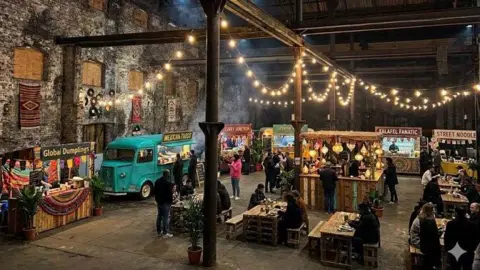 John Taylor Architect Artist's impression of the interior of the Sugar Sheds, with food trucks and pop-up stalls, along with lighting hanging down from the building.