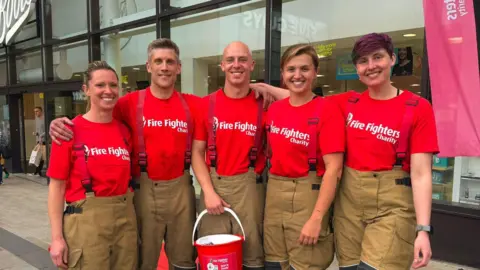 Royal Berkshire Fire and Rescue Service A group of firefighters - two men and three women - pose for a photo. They are holding a charity bucket and wearing red T-shirts with Fire Fighters Charity printed on the front in white letters.