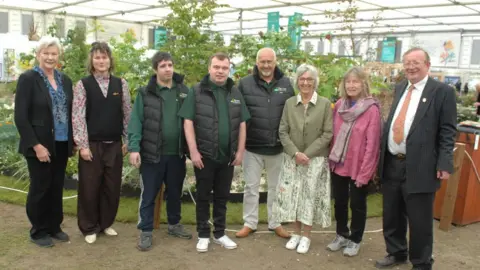 A group photo of two members of Grow standing alongside six members of the Cornish-based Burncoose Nurseries. In the background is the garden.