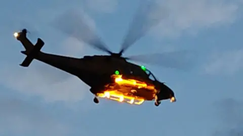 A helicopter in mid-air, seen from the ground and silhoutted against a blue sky with fading light.