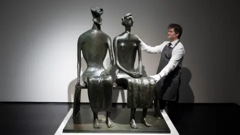 A bronze sculpture of a couple sitting on a bench. A man in white shirt and apron poses with his hands on the sculpture
