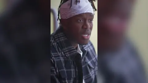 KSI is wearing a white bandana and a blue shirt. He looks off camera.