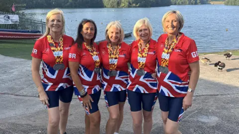 Five women, who are wearing red Team GB T-shirts and medals around their necks, are grouped together for a photograph in front of the water. 