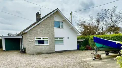 A detached house with an attached garage and gravel drive. The left hand side of the building has beige exposed brick, and the right hand side of the building is painted white. A green and blue boat can be seen in the driveway. 