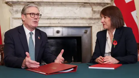 Keir Starmer and Rachel Reeves sit facing each other inside Downing Street, with the red Budget folder on the desk in front of them and the Union flag behind them, in October