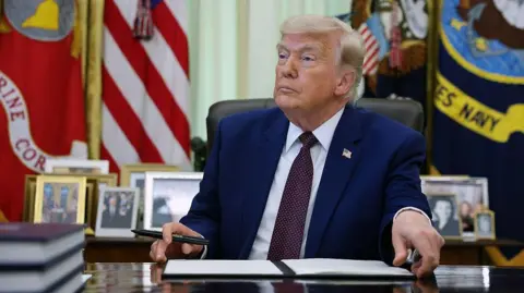 Donald Trump sits in the Oval Office with his hands holding a notepad of paper and a pen. He is wearing a navy suit and burgundy tie. 