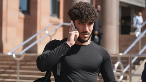 PA Media Mazyar Azarbonyad leaves Newcastle Crown Court wearing a black, fitted long-sleeved top. He is holding a mobile phone to his ear as he looks ahead.