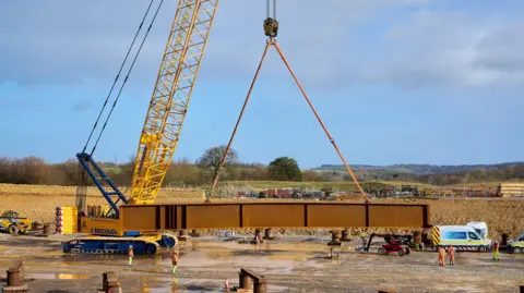 National Highways A yellow and blue crane holding up a large copper coloured beam which is suspended in mid air. Below it are five workers, dressed in orange and yellow hi-vis, who are navigating the lowering of the beam. It is a sunny day with blue skies and wispy clouds. 