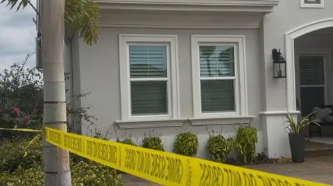Manatee County Sheriff’s Office A grey/cream Florida home, with a palm tree in the foreground. There is yellow police tape wrapped around the tree. It's a close up picture of the house, showing two front facing windows, with a series of plants running around the front of the house.