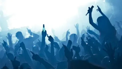 Ravers crowd in front of a DJ on a stage. Some of the dancers are on people's shoulders. Some are holding bottles in the air. They are silhouetted by the glaring lights. The picture has ben taken from behind the dancers.