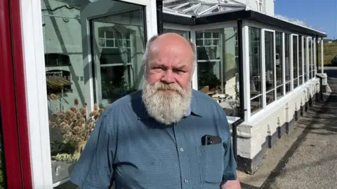BBC/Phil Harrison Philip Harris, a middle aged man with a bushy grey beard, wears a blue sleeveless shirt and stands outside the white-fronted Sportsman pub in Seasalter.