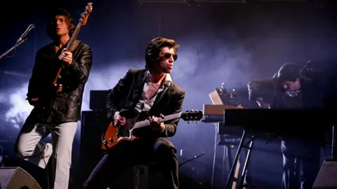 Three members of Arctic Monkeys on a smoky stage at Glastonbury at night. On the left, a man plays a bass guitar, the neck raised towards the sky. He has bushy brown hair and wears a black leather jacket and light-grey trousers. In the centre, a man with brown hair plays a brown guitar while looking to his left. He wears sunglasses, a dark suit and white shirt, unbuttoned at the chest. To the right, a man with short hair bends over a keyboard, his right hand on the keys. 
