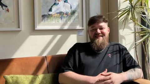 Harriet Heywood/BBC Mr Kabzinski is sitting on a brown couch, with green pillows, next to a window on a sunny day. He is wearing a black top and silver chain. His hands are clasped in front of him. Mr Kabzinski has brown hair, a long beard and is smiling at the camera.