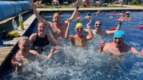BBC Six swimmers, a mixture of men and women, who are taking part in the challenge, are in the pool posing for the camera. For most, their hands are in the air and they are smiling.