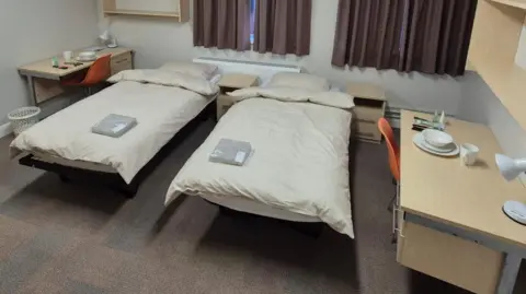 West Northamptonshire Council A room with two beds in it. They have cream covers. On the right is a desk with crockery and a lamp on it. The floor is brown and there are brown curtains. There are shelves are on the walls.