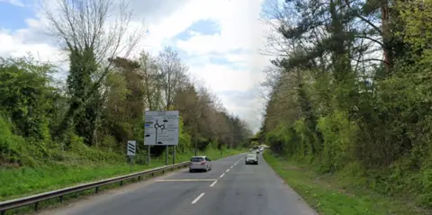 Google Google maps image of the A4501 road between B&Q and Aldi in Cwmbran Drive, Cwmbran. Roundabout signage can be seen on the left hand side of the road. Green grass and trees line either side of a single carriageway. A silver car is driving away on the left hand side of the road, while a line of cars travels towards the camera on the right. 