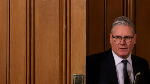 Keir Starmer arrives for a meeting with leaders from across society to discuss tackling antisemitism, at Downing Street in London in May. He is wearing a dark suit, white shirt and tie, and is pictured appearing round a door.