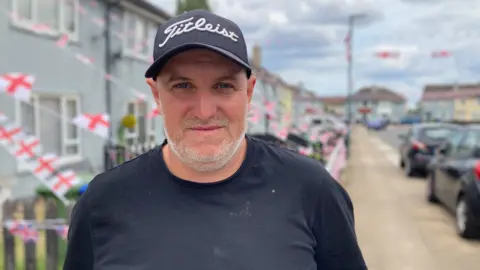 Simon Spark / BBC Tom Leonard standing in front of the street filled with flags. He has a grey beard and wears a black baseball cap and shirt