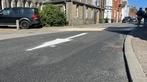 Andrew Turner/BBC Fresh asphalt laid on Deneside in Great Yarmouth, with a newly-painted white arrow road marking in the centre of the image denoting the change in direction of the one-way system. Behind the road is the side of Christchurch and other buildings on Deneside, with some vehicles parked.