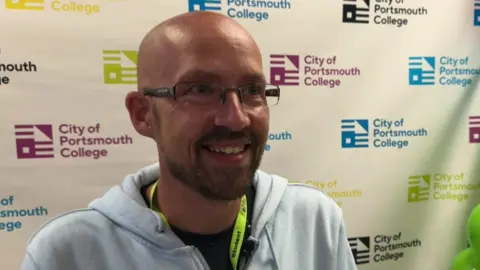 BBC A bald man wearing glasses is grinning. He is standing in front of a screen which has the City of College Portsmouth logo on.