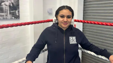 Serena is standing in a boxing ring. She is wearing a blue hoodie which says Empire Fighting Chance on the front. 