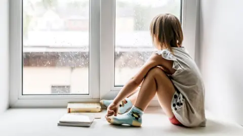 A child sits on a window sill looking out on a rainy day. 