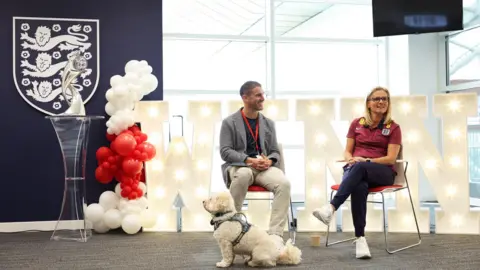 Getty Images Gavin Step sits next to Lionesses manager Sarina Wiegman at an event. The Women's Euros trophy is on a plinth next to them. A small, white dog is sitting at their feet and there are red and white balloons to the side of the picture. 