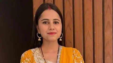 A woman in a yellow and white trimmed traditional Indian saree holds her phone in her left hand standing by a wooden doorway.